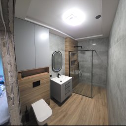 Lux-bud - Nowoczesna łazienka po remoncie: szare płytki imitujące beton, drewnopodobne akcenty, kabina prysznicowa typu walk-in i podświetlane lustro nad umywalką.