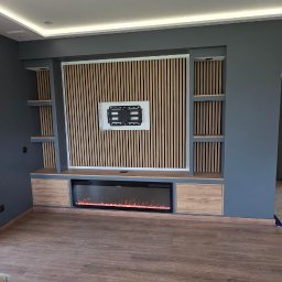 Lux-bud - Nowoczesny salon z szarą ścianą i drewnianą zabudową TV z elektrycznym kominkiem. Widoczne schody w tle i duże okno z widokiem na zieleń.