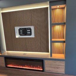 Lux-bud - Nowoczesna ściana TV z drewnianymi lamelami, elektrycznym kominkiem i podświetlanymi półkami w szaro-beżowej tonacji. Schody w tle w trakcie wykończenia.