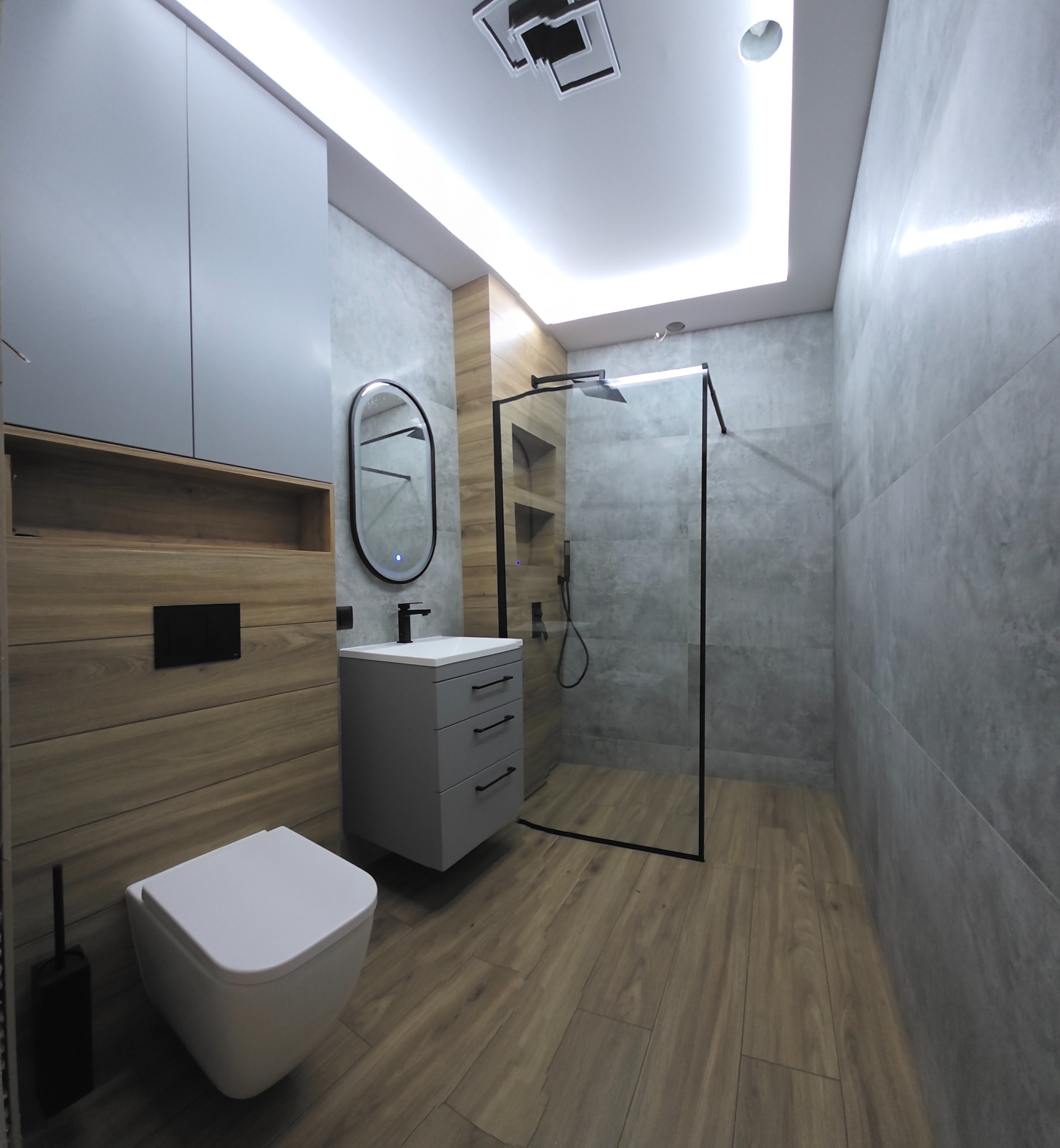 Nowoczesna łazienka z drewnopodobną podłogą i szarą armaturą. Kabina prysznicowa typu walk-in z czarnymi ramami. Lustro owalne z podświetleniem LED.