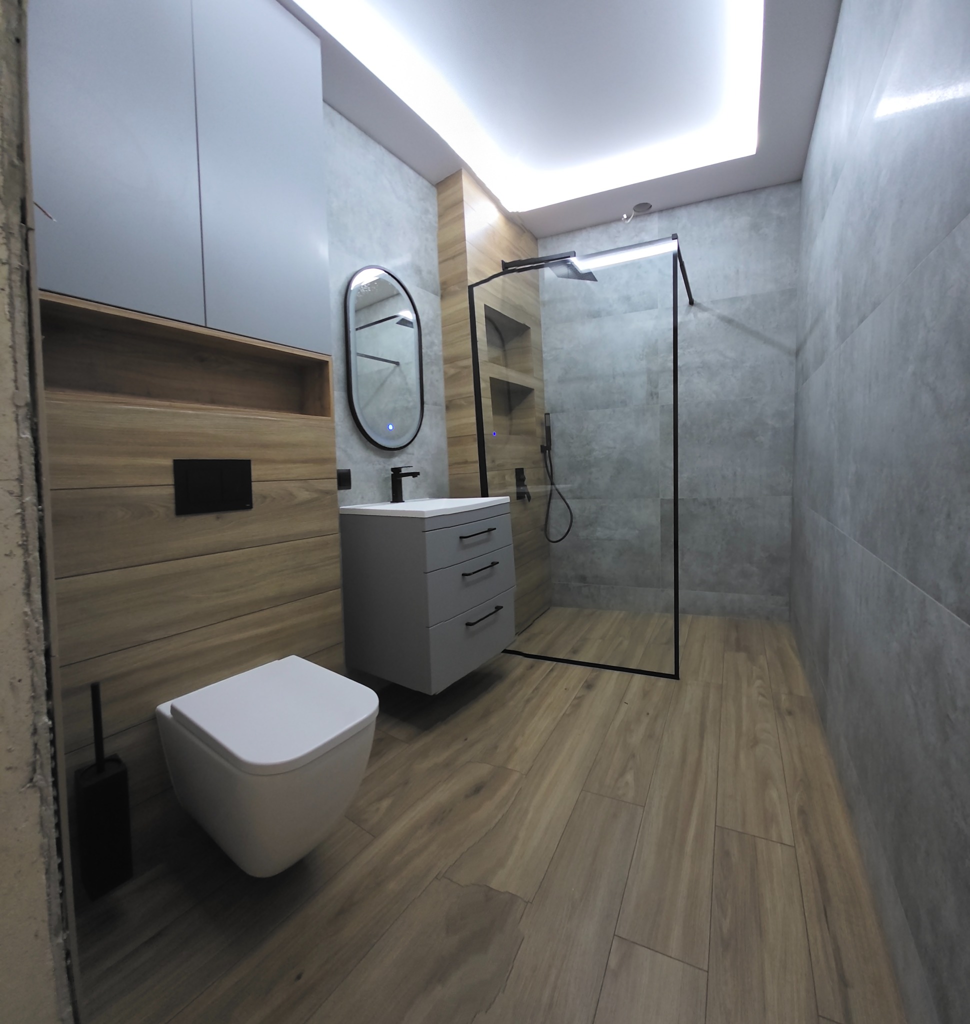 Nowoczesna łazienka w szarościach i drewnie: kabina walk-in z czarnymi ramami, podwieszana toaleta, szafka z umywalką i lustro z podświetleniem LED.