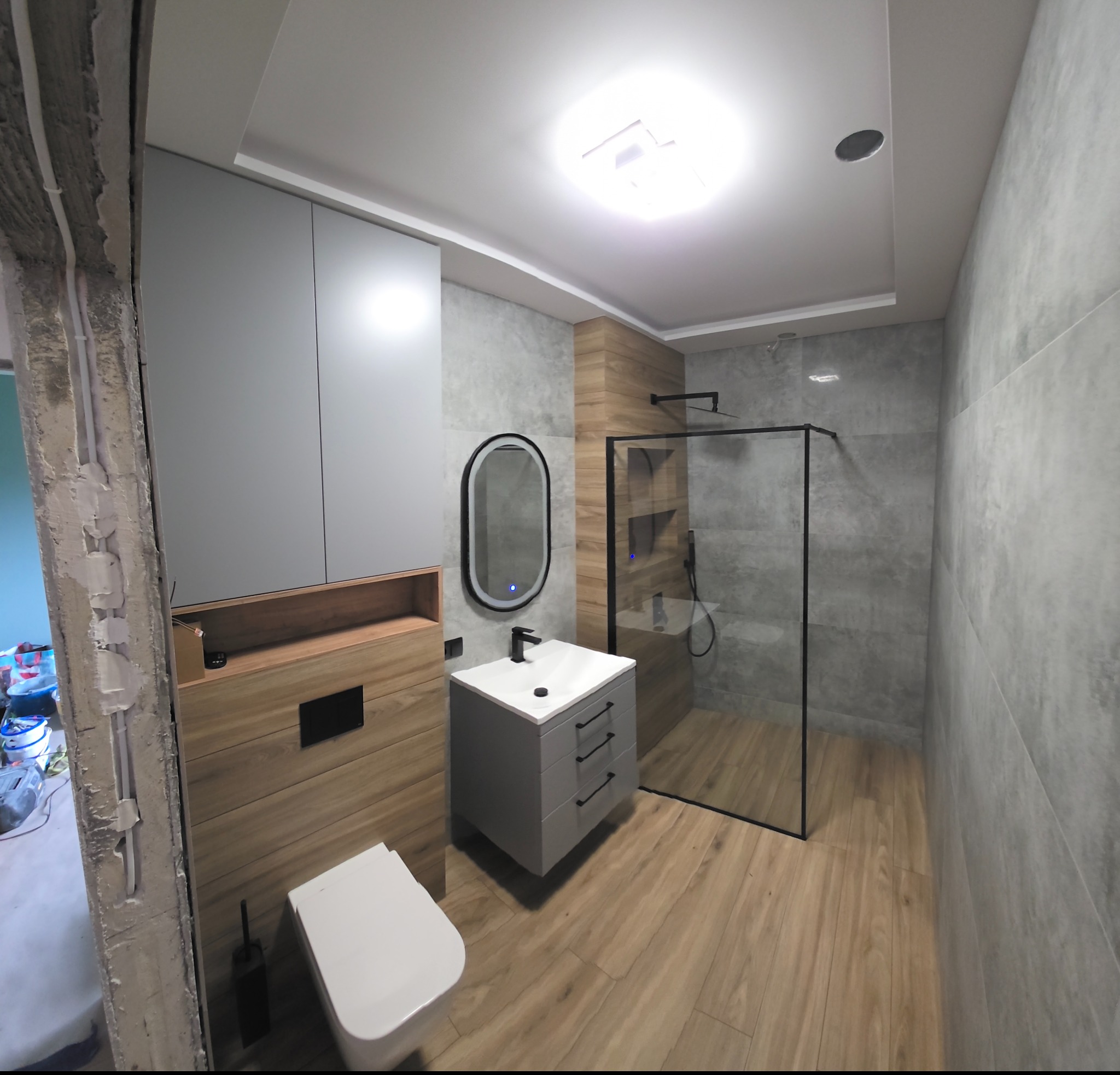 Nowoczesna łazienka po remoncie: szare płytki imitujące beton, drewnopodobne akcenty, kabina prysznicowa typu walk-in i podświetlane lustro nad umywalką.