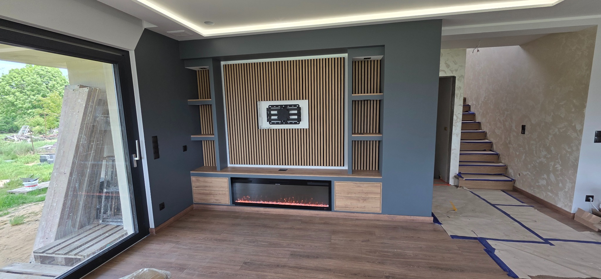 Nowoczesny salon z szarą ścianą i drewnianą zabudową TV z elektrycznym kominkiem. Widoczne schody w tle i duże okno z widokiem na zieleń.