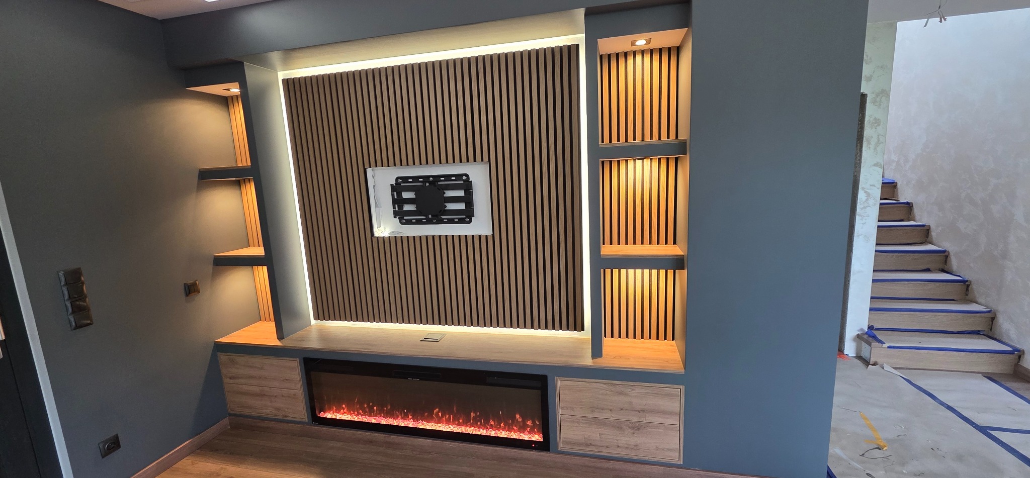 Nowoczesna ściana TV z drewnianymi lamelami, elektrycznym kominkiem i podświetlanymi półkami w szaro-beżowej tonacji. Schody w tle w trakcie wykończenia.