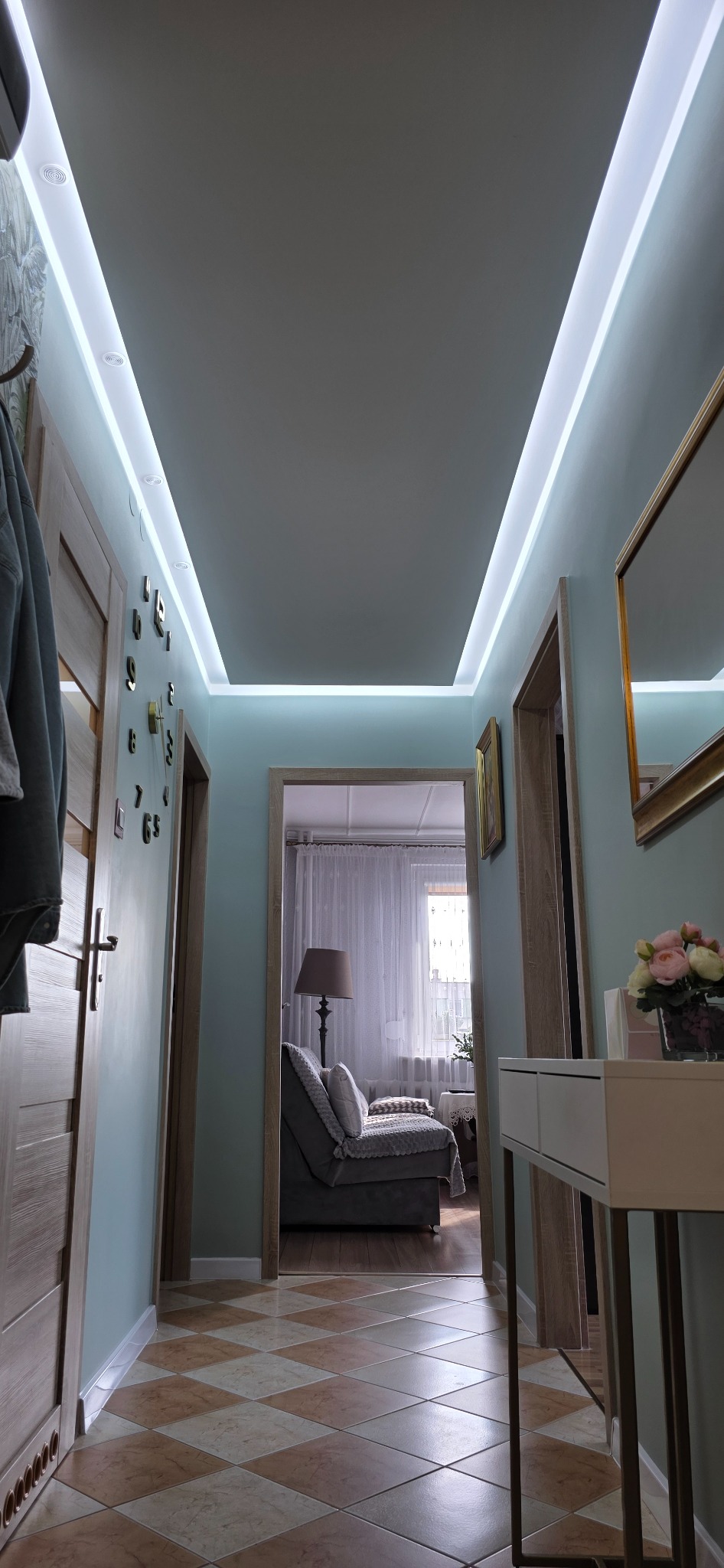 Wnętrze korytarza z oświetleniem LED na suficie, widoczne drzwi, zegar na ścianie i fragment pokoju z sofą w tle. Podłoga wyłożona płytkami w romby.