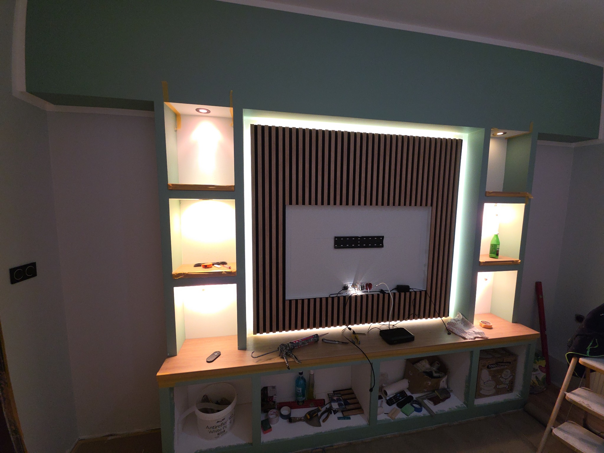 Wykończona ściana TV z drewnianymi lamelami, podświetleniem LED i półkami. Widoczne narzędzia i materiały budowlane, sugerujące etap prac wykończeniowych.