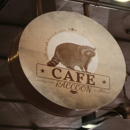 Piotr Dąbrowski - Okrągły szyld kawiarni 'Cafe Raccoon' z wizerunkiem szopa, zawieszony pod sufitem. Styl vintage, brązowe barwy, rok założenia 1973.