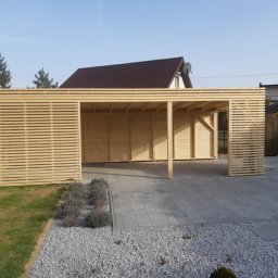 Paweł Szopiński - Nowoczesna, drewniana wiata garażowa z lamelami,  wykończona kostką brukową i żwirem, otoczona zielenią. Widok na dom z brązowym dachem w tle.