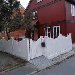 Paweł Szopiński - Białe, dekoracyjne ogrodzenie posesji z furtką i skrzynką na listy, na tle czerwonego, dwukondygnacyjnego domu z białymi oknami. Widok z ulicy.