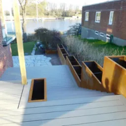 Nowoczesny taras z szarych desek kompozytowych z metalowymi donicami schodkowymi. Widok na jezioro i ceglany budynek w tle, podkreślający unikalny design.