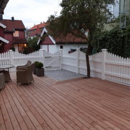 Paweł Szopiński - Drewniany taras z białym płotem na tle domów. W tle widoczna zieleń. Na tarasie stolik i krzesła z rattanu.