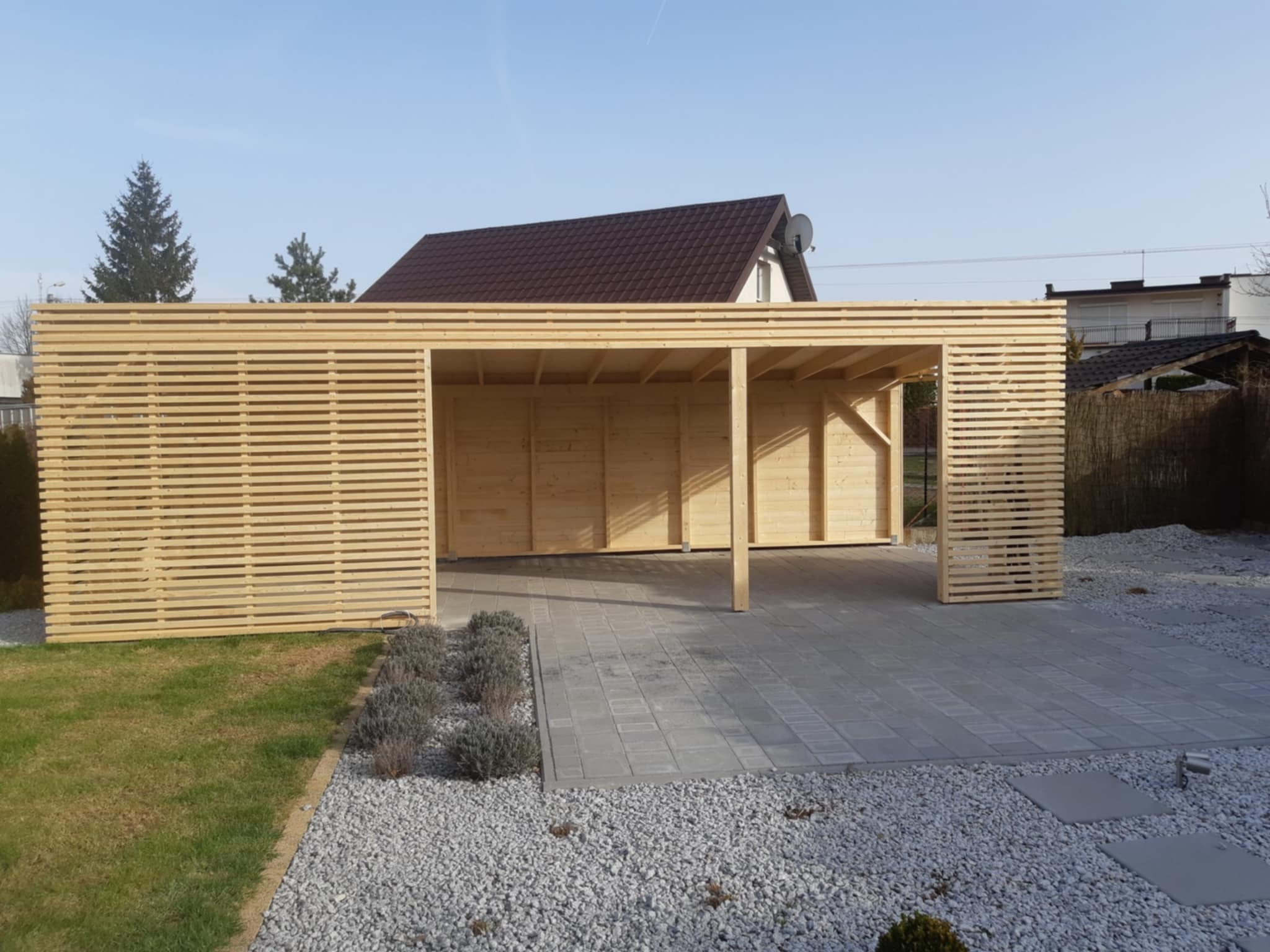 Nowoczesna, drewniana wiata garażowa z lamelami,  wykończona kostką brukową i żwirem, otoczona zielenią. Widok na dom z brązowym dachem w tle.