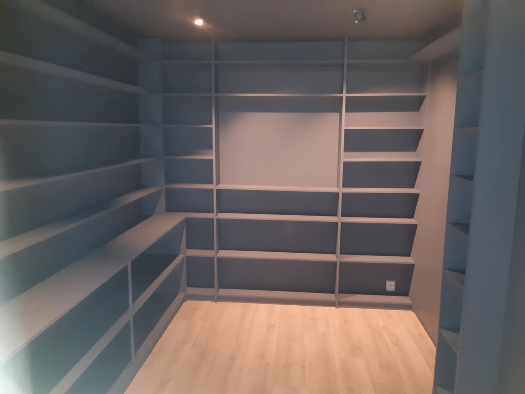 Szara zabudowa meblowa na wymiar z półkami, zintegrowana ze ścianą, na drewnianej podłodze. Widoczne gniazdko elektryczne w dolnej części zabudowy.