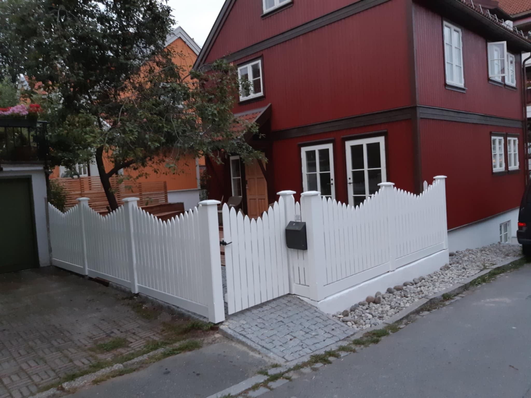 Białe, dekoracyjne ogrodzenie posesji z furtką i skrzynką na listy, na tle czerwonego, dwukondygnacyjnego domu z białymi oknami. Widok z ulicy.