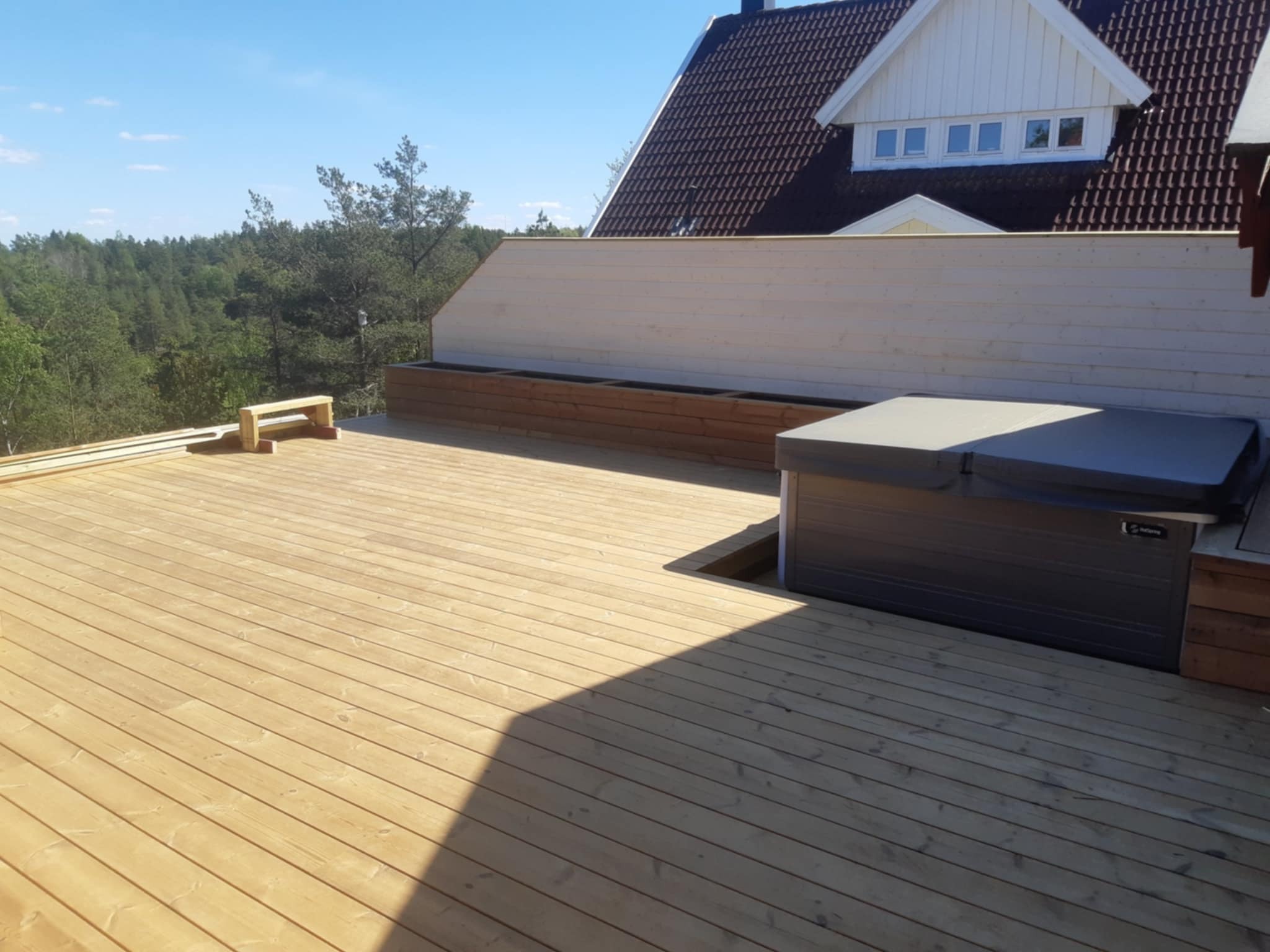 Jasna, drewniana podłoga tarasu z jacuzzi wbudowanym w konstrukcję. W tle widoczny las i fragment domu z brązowym dachem.