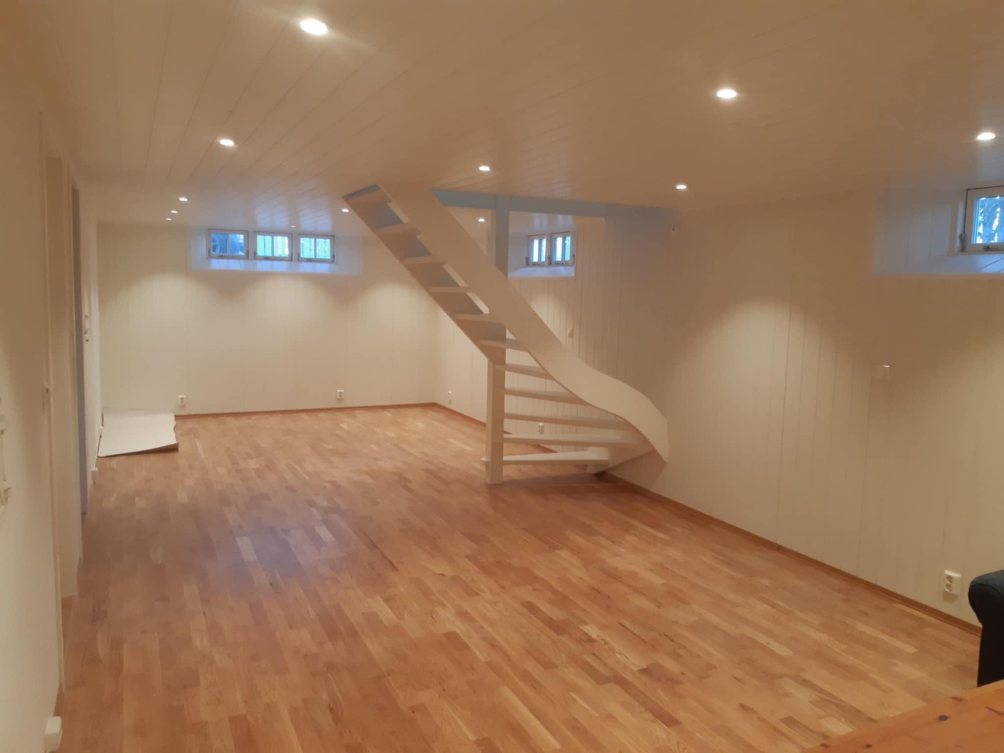 Jasne drewniane panele podłogowe w przestronnym, odnowionym wnętrzu z białymi ścianami i oryginalnymi, spiralnymi schodami. Widoczne okna piwniczne i punktowe oświetlenie.