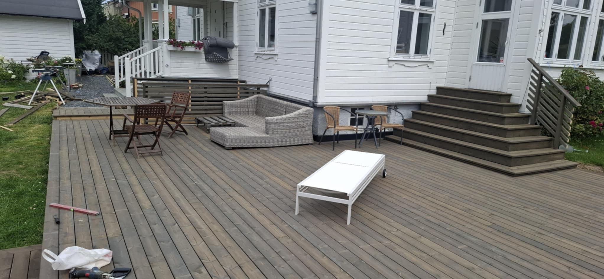 Drewniany taras z meblami ogrodowymi i schodami przy białym domu. W tle widoczne narzędzia i materiały budowlane, sugerujące trwające prace wykończeniowe.