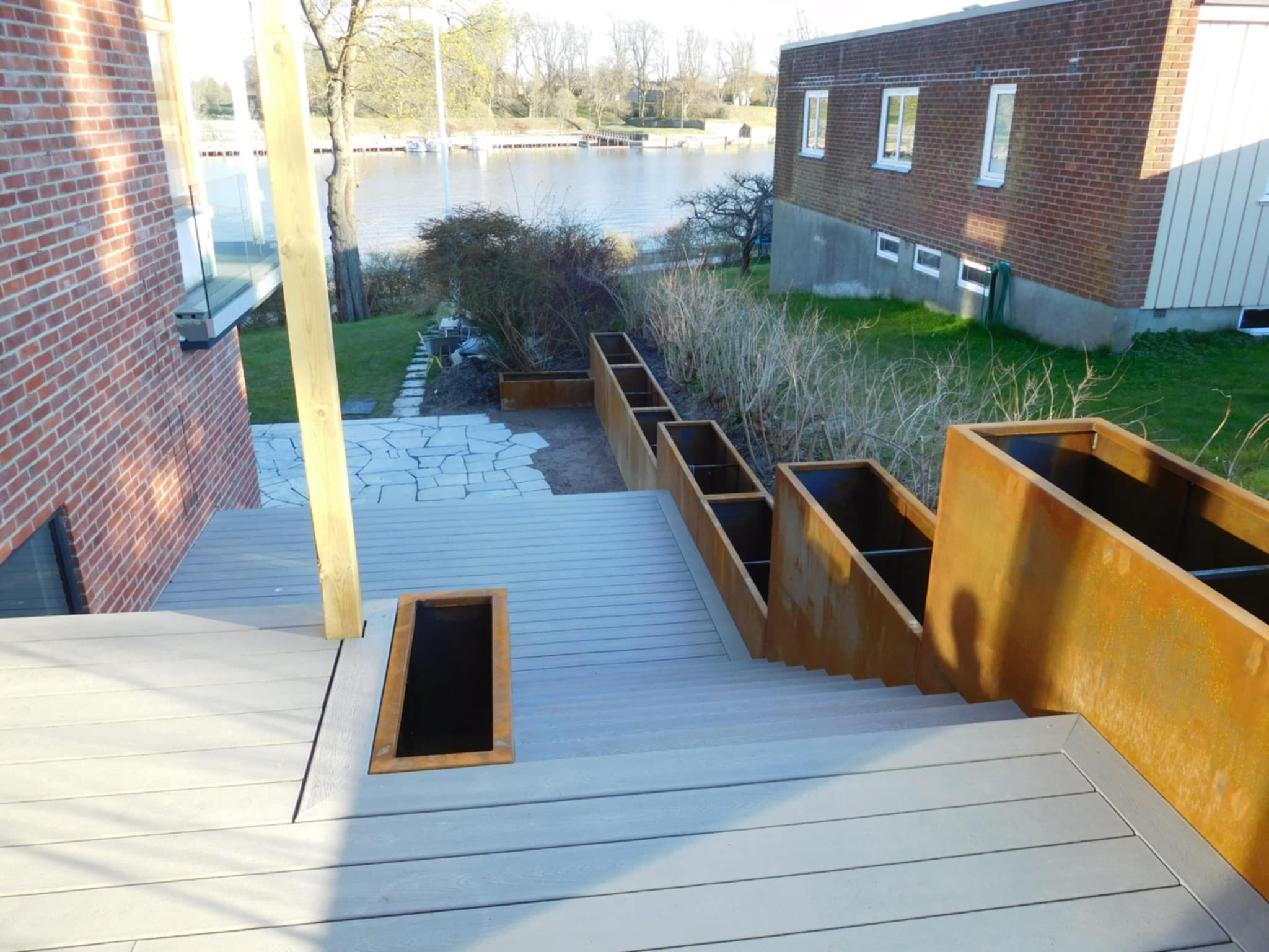 Nowoczesny taras z szarych desek kompozytowych z metalowymi donicami schodkowymi. Widok na jezioro i ceglany budynek w tle, podkreślający unikalny design.