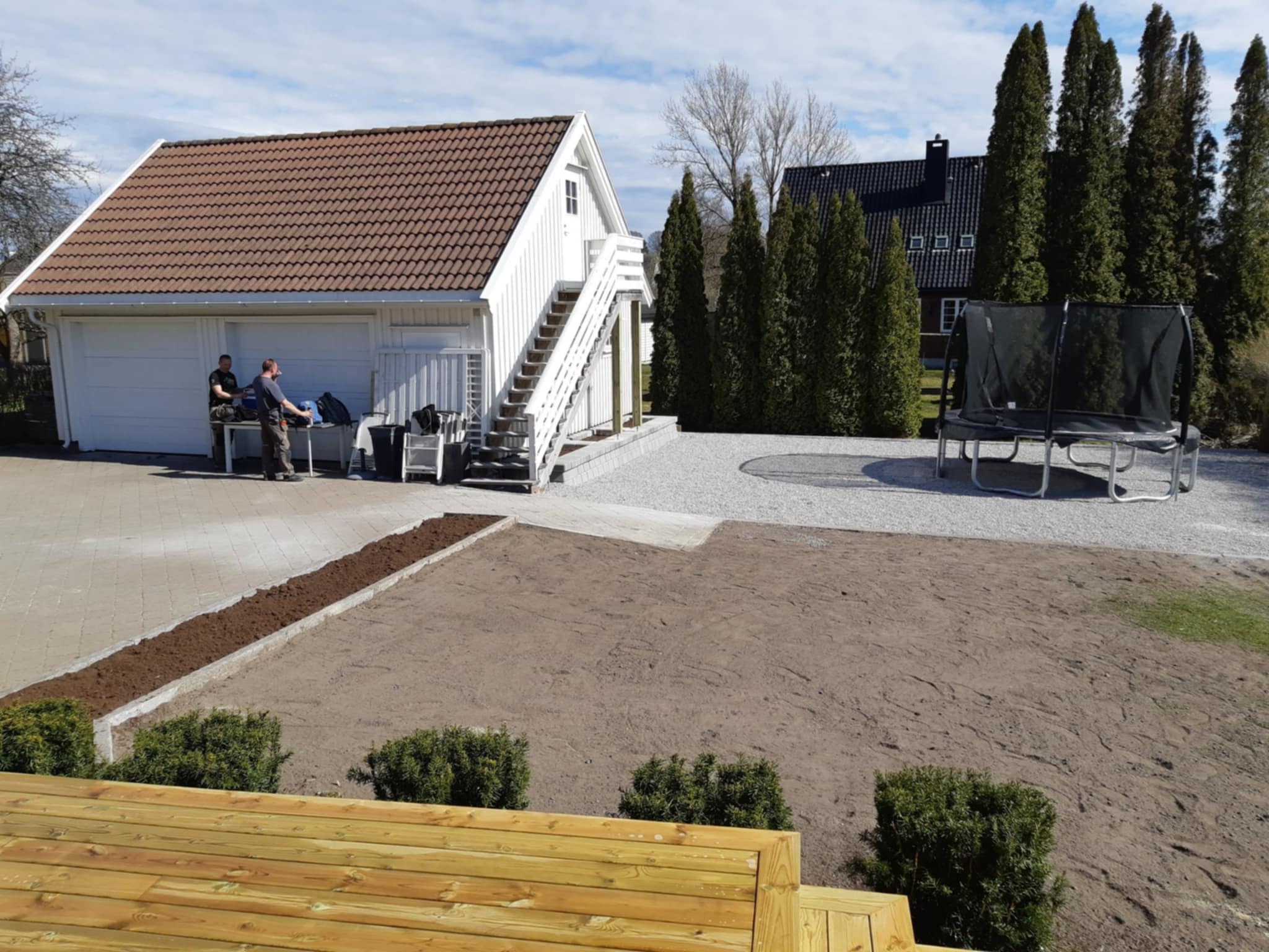 Przygotowany teren pod trawnik z obrzeżami, widoczne budynki gospodarcze i trampolina. Dwóch mężczyzn przy pracy na zewnątrz.