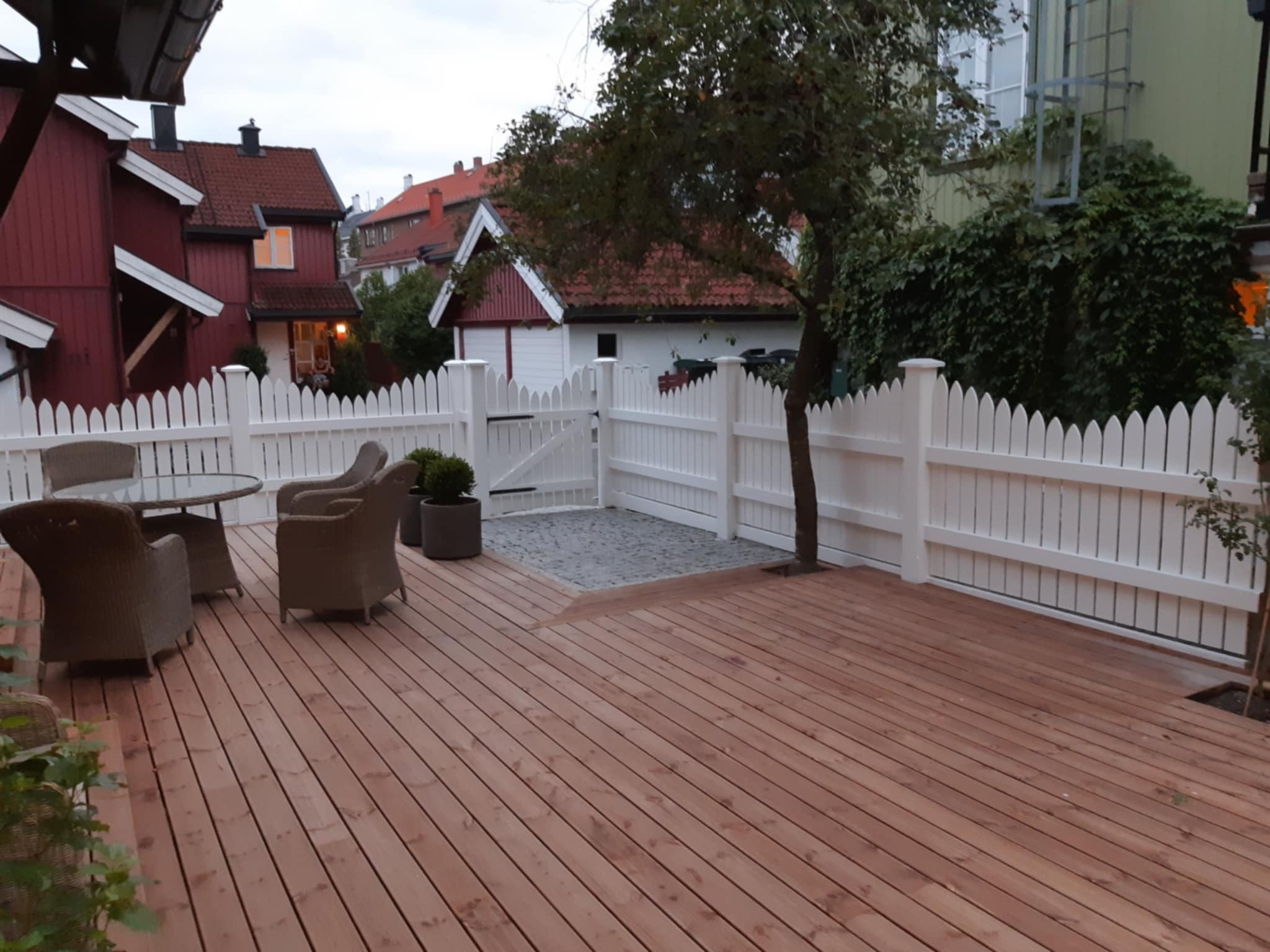 Drewniany taras z białym płotem na tle domów. W tle widoczna zieleń. Na tarasie stolik i krzesła z rattanu.