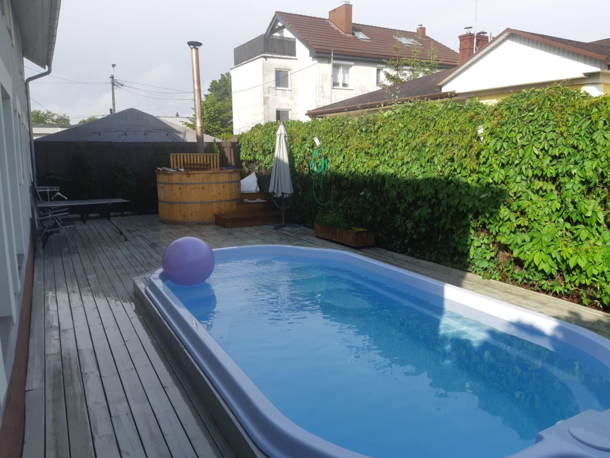 Drewniany taras z basenem i balią. W tle zielony żywopłot i budynki mieszkalne. Na tarasie leży fioletowa piłka. Balia z kominem.