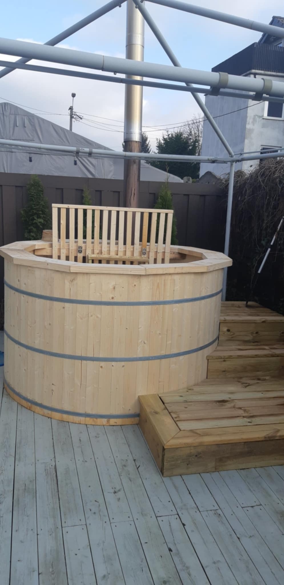 Drewniana balia ogrodowa z piecem zewnętrznym i schodkami, umieszczona na tarasie z desek. Widoczne elementy konstrukcji zadaszenia i otoczenie ogrodu.