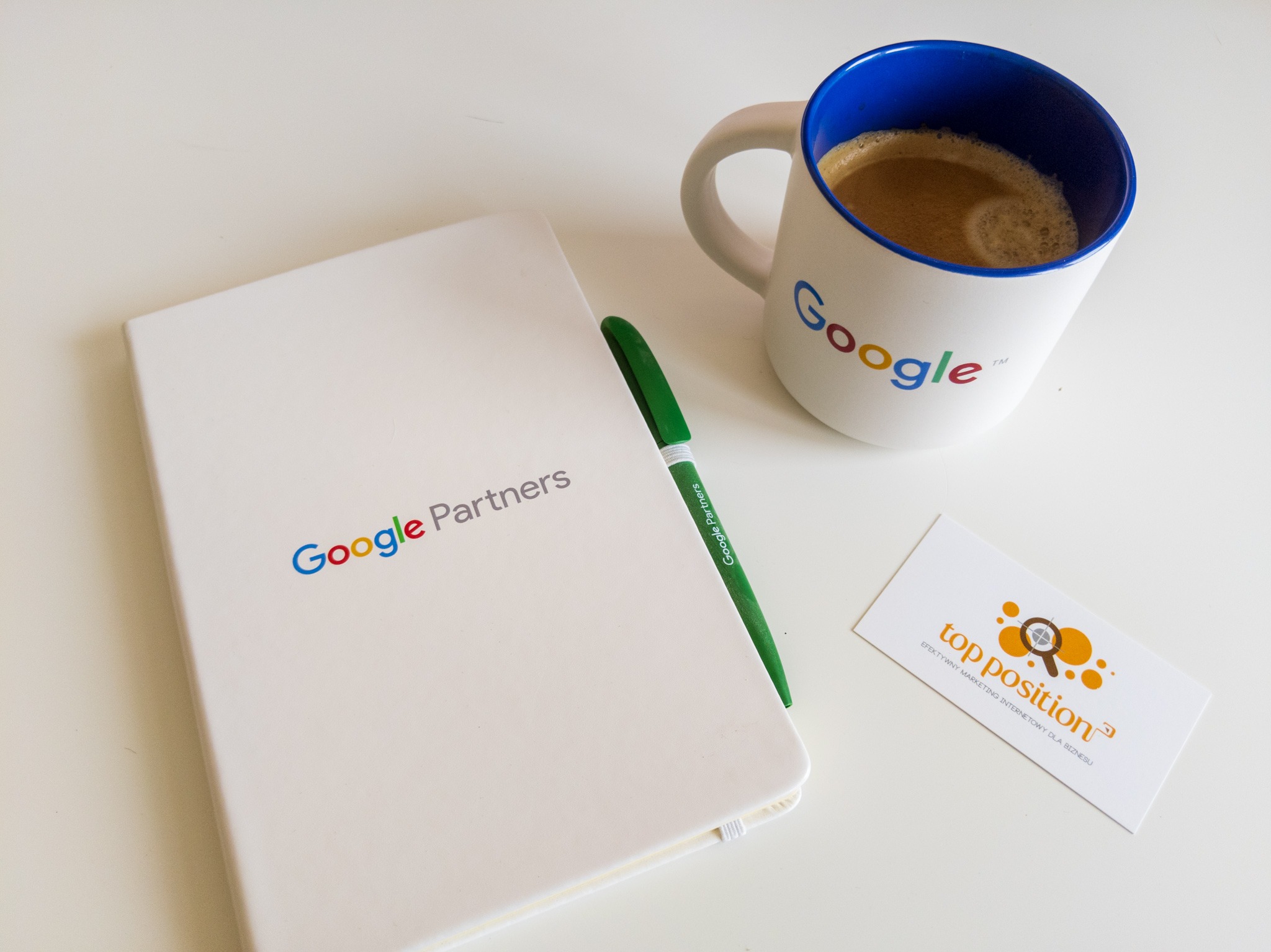 Biurko z notatnikiem Google Partners, długopisem, wizytówką z napisem 'top position' i kubkiem kawy z logo Google na białym tle.