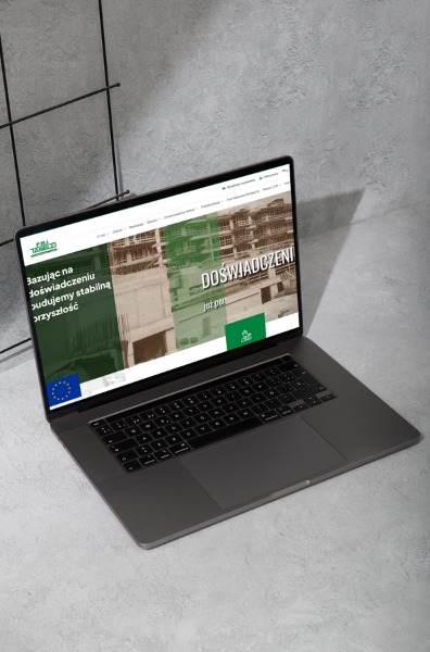 Laptop wyświetla stronę internetową fbitasbud.pl z zielonym logotypem firmy na tle surowego, szarego betonu i metalowej kratki.