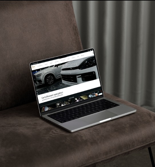 Laptop z wyświetloną stroną internetową custommadestudio.pl, prezentującą zdjęcia samochodów, umieszczony na brązowym fotelu.