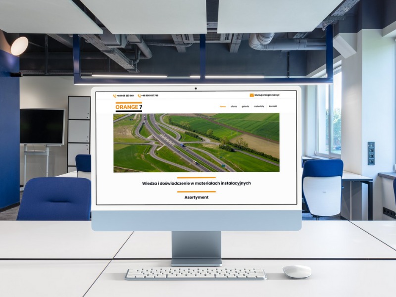 Monitor wyświetla stronę internetową orangeseven.pl z widokiem na autostradę z lotu ptaka, umieszczony na biurku w nowoczesnym biurze.