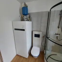 Nowoczesna łazienka z białą pompą ciepła Panasonic, toaletą z zabudowaną spłuczką i kabiną prysznicową z czarnymi elementami. Całość w jasnych barwach.