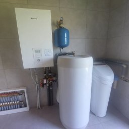 Budo-Therm Wojciech Kłos - Biały piec Panasonic Aquarea z niebieskim zbiornikiem wyrównawczym, obok biały zmiękczacz wody i rozdzielacz podłogowy w kotłowni wykończonej płytkami.