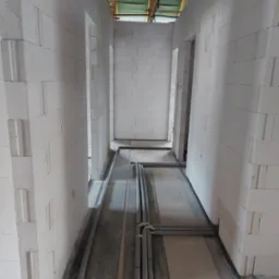 Wnętrze w trakcie budowy: białe ściany z betonu komórkowego, szare rury instalacyjne ułożone na podłodze, surowy strop z drewnianych belek.