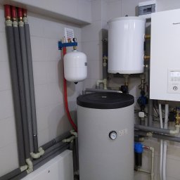Budo-Therm Wojciech Kłos - Biała instalacja grzewcza Panasonic z zbiornikiem i rurami w szarych otulinach, zamontowana na tle białych płytek. Widoczny manometr i elementy hydrauliczne.