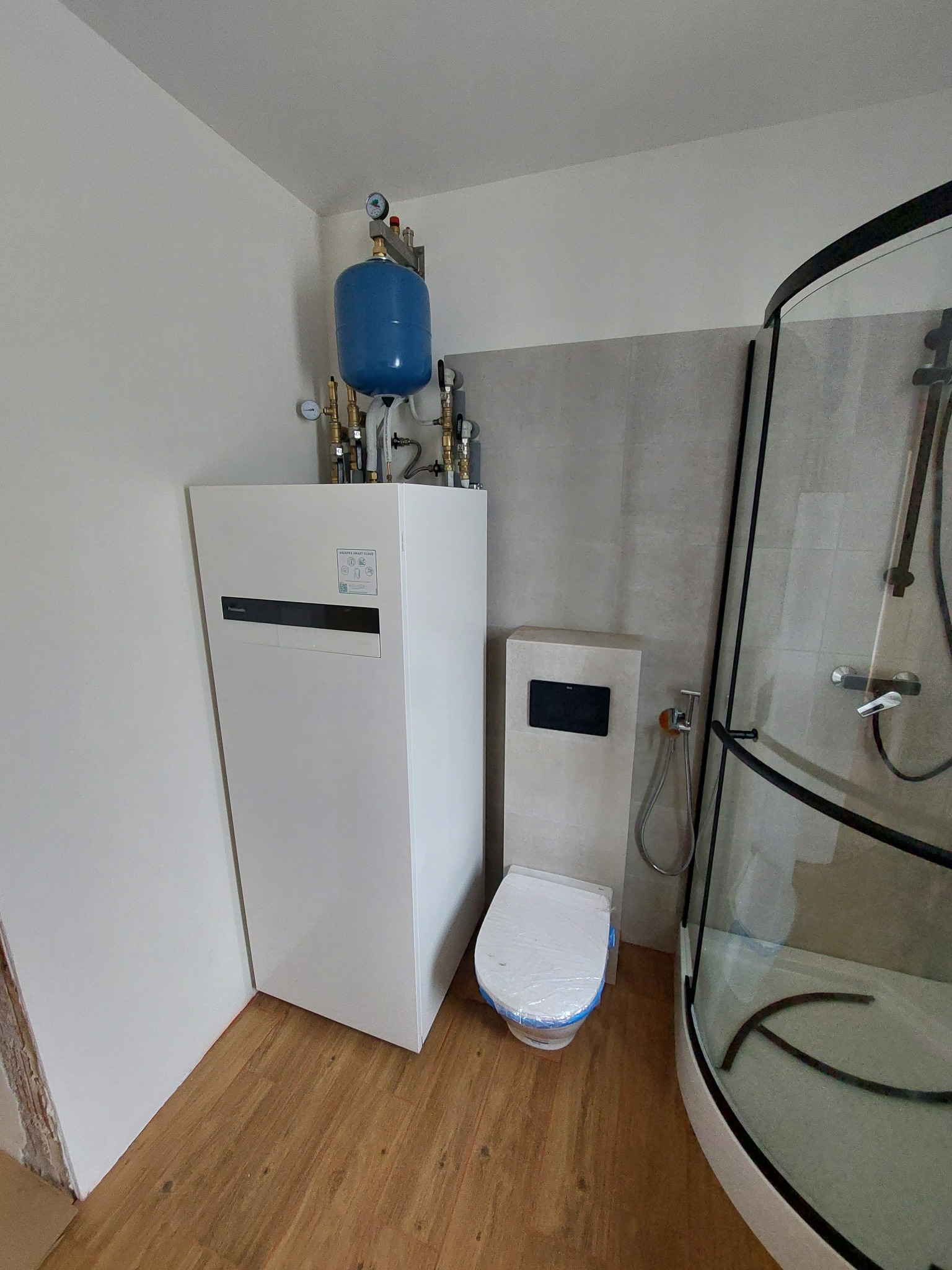 Nowoczesna łazienka z białą pompą ciepła Panasonic, toaletą z zabudowaną spłuczką i kabiną prysznicową z czarnymi elementami. Całość w jasnych barwach.