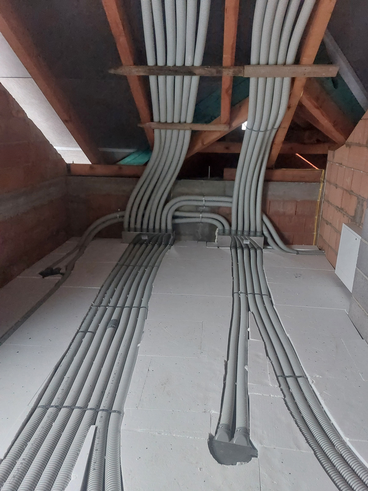 Szare rury karbowane ułożone równolegle w wyżłobieniach poddasza, przymocowane do drewnianej konstrukcji. Widoczne połączenia i fragment ściany z cegły.