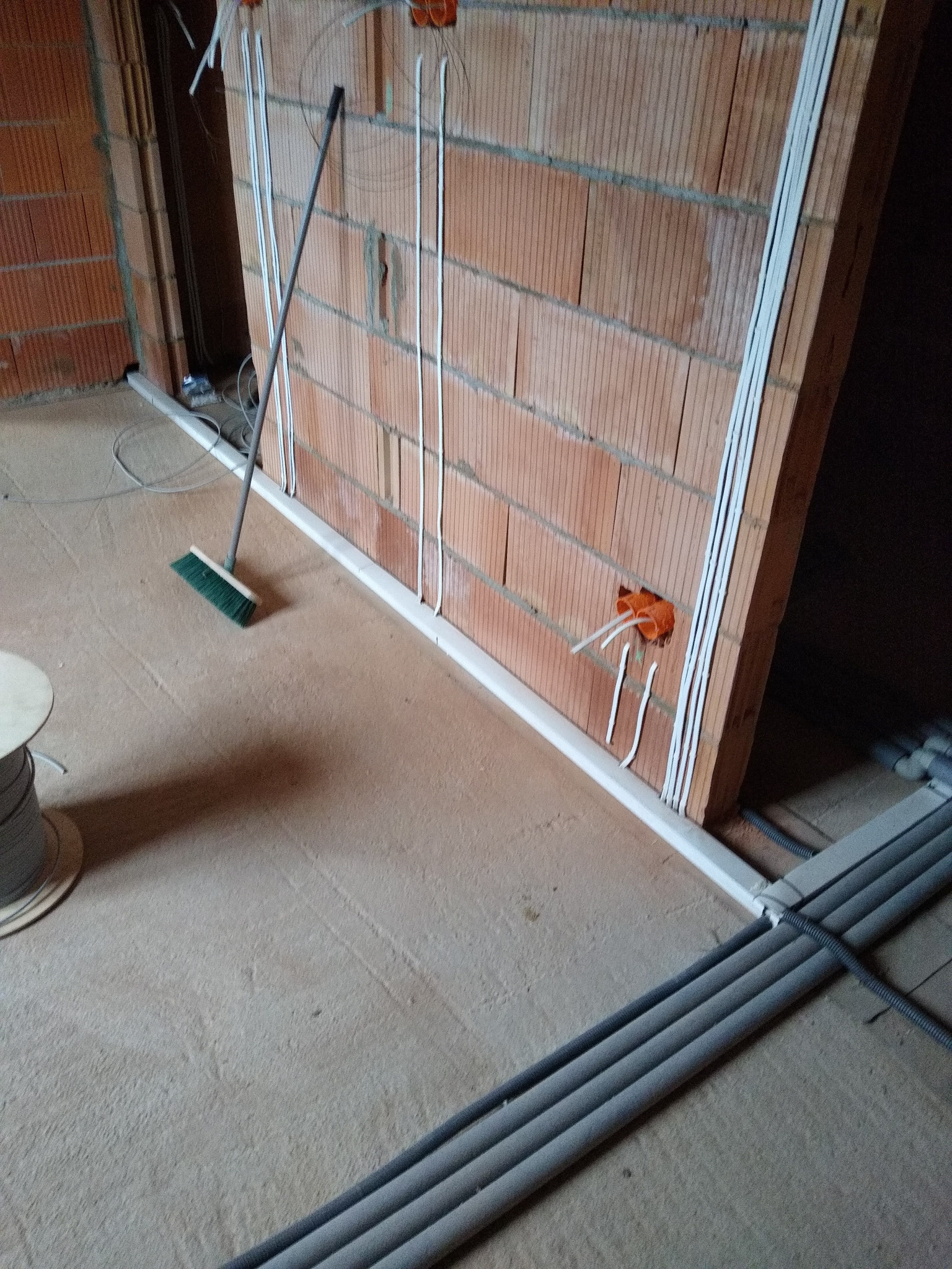Instalacja elektryczna w trakcie montażu: przewody w białych listwach na ceglanej ścianie, puszki instalacyjne, szara rura karbowana na podłodze, widok z perspektywy podłogi.