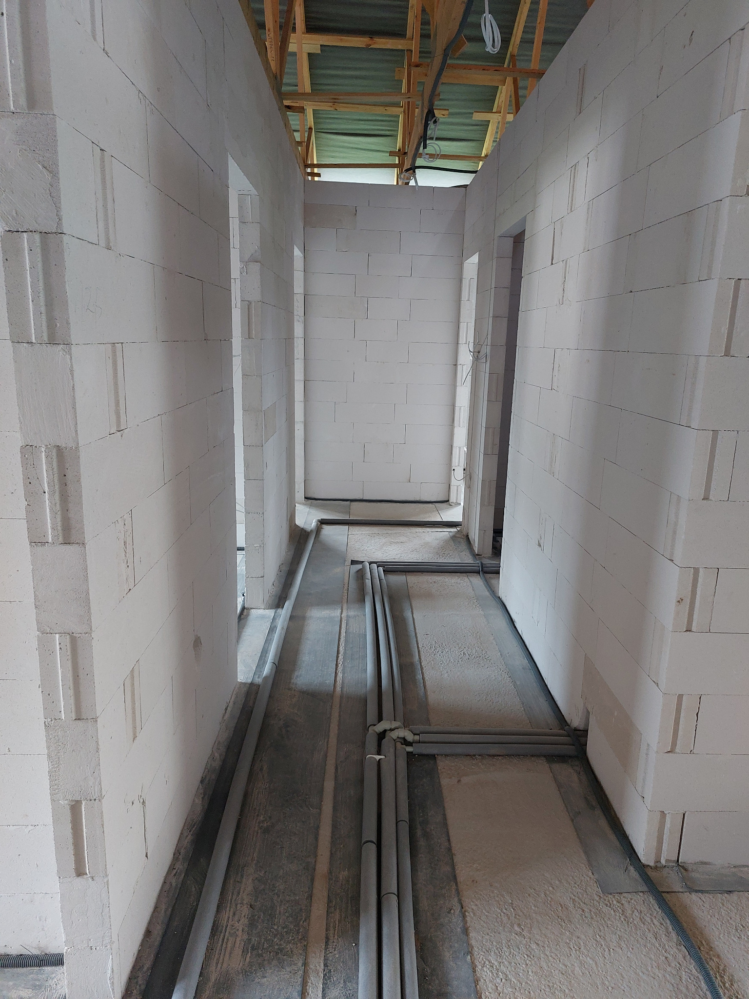 Wnętrze w trakcie budowy: białe ściany z betonu komórkowego, szare rury instalacyjne ułożone na podłodze, surowy strop z drewnianych belek.