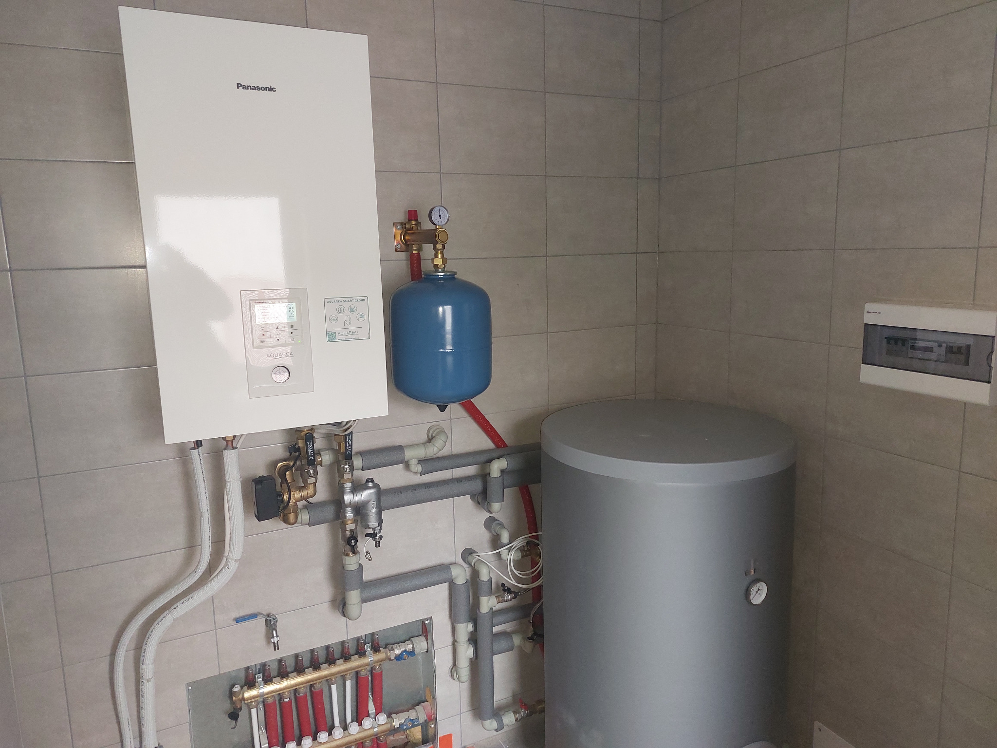 Biały piec gazowy Panasonic z niebieskim zbiornikiem wyrównawczym i szarym zasobnikiem ciepłej wody, na tle kafelkowanej ściany w kotłowni. Widoczne rury instalacji CO.
