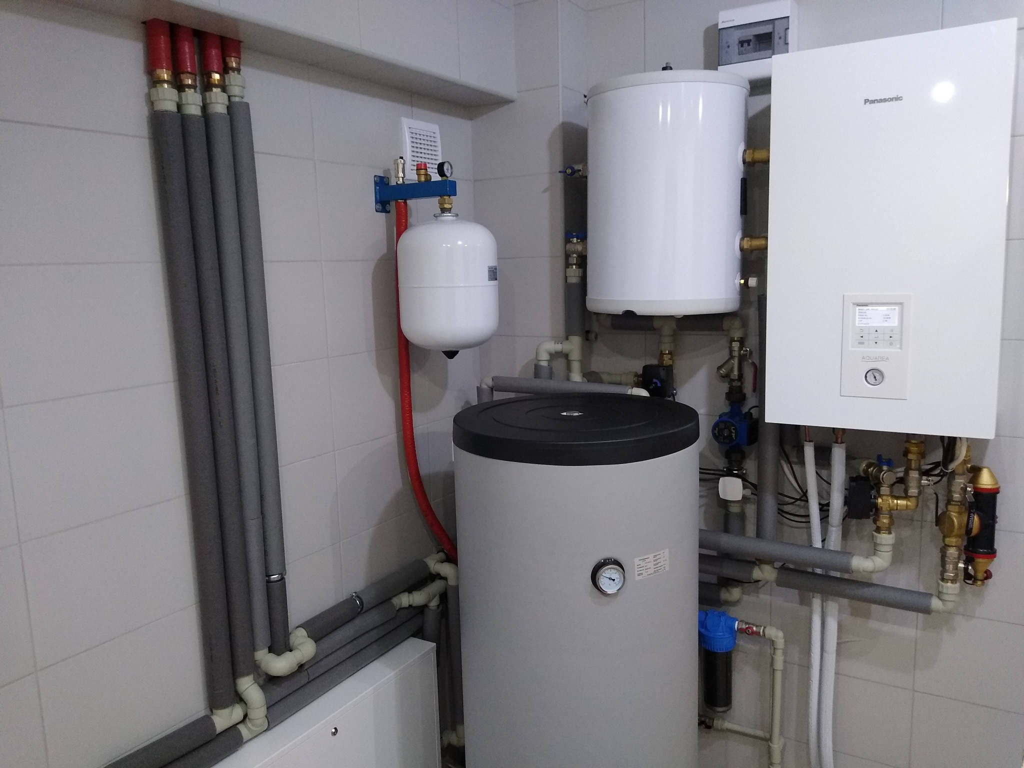 Biała instalacja grzewcza Panasonic z zbiornikiem i rurami w szarych otulinach, zamontowana na tle białych płytek. Widoczny manometr i elementy hydrauliczne.