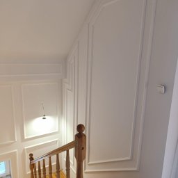 SOLID Jakub Ragus - Klatka schodowa z drewnianymi schodami i białymi ścianami zdobionymi sztukaterią. Widoczne detale wykończenia, oświetlenie i fragment okna.