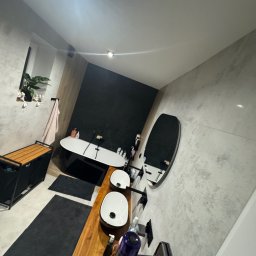 SOLID Jakub Ragus - Nowoczesna łazienka z owalną wanną, drewnianym blatem pod umywalki i okrągłym lustrem. Szaro-białe płytki na ścianach, czarne akcenty i roślina w oknie.