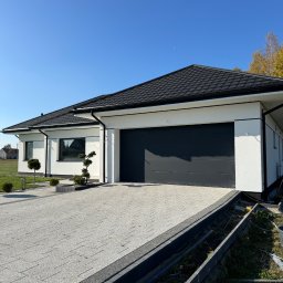 SOLID Jakub Ragus - Nowoczesny dom jednorodzinny z grafitową bramą garażową i kostką brukową na podjeździe w słoneczny dzień. Widoczne detale wykończenia elewacji i zagospodarowania terenu.
