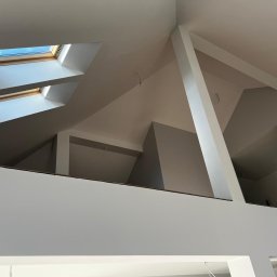 SOLID Jakub Ragus - Poddasze ze skosami i oknami dachowymi po malowaniu na biało i szaro, widoczne elementy konstrukcyjne i fragment ściany. Oświetlenie naturalne.