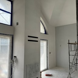 SOLID Jakub Ragus - Wnętrze po malowaniu w Żelechowie: ściany na biało, okna zabezpieczone folią, widoczne rusztowania. Minimalistyczny design z elementami surowego wykończenia.