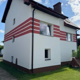 Dwupiętrowy dom z białą elewacją i czerwonymi pasami, brązowym dachem i rynnami. Widok na fragment ogrodu i niebo z chmurami.