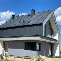 SOLID Jakub Ragus - Nowoczesny dom z szarą blachą na dachu i oknami dachowymi, elewacja w trakcie wykończenia, widoczne elementy rusztowania i składowane materiały budowlane.