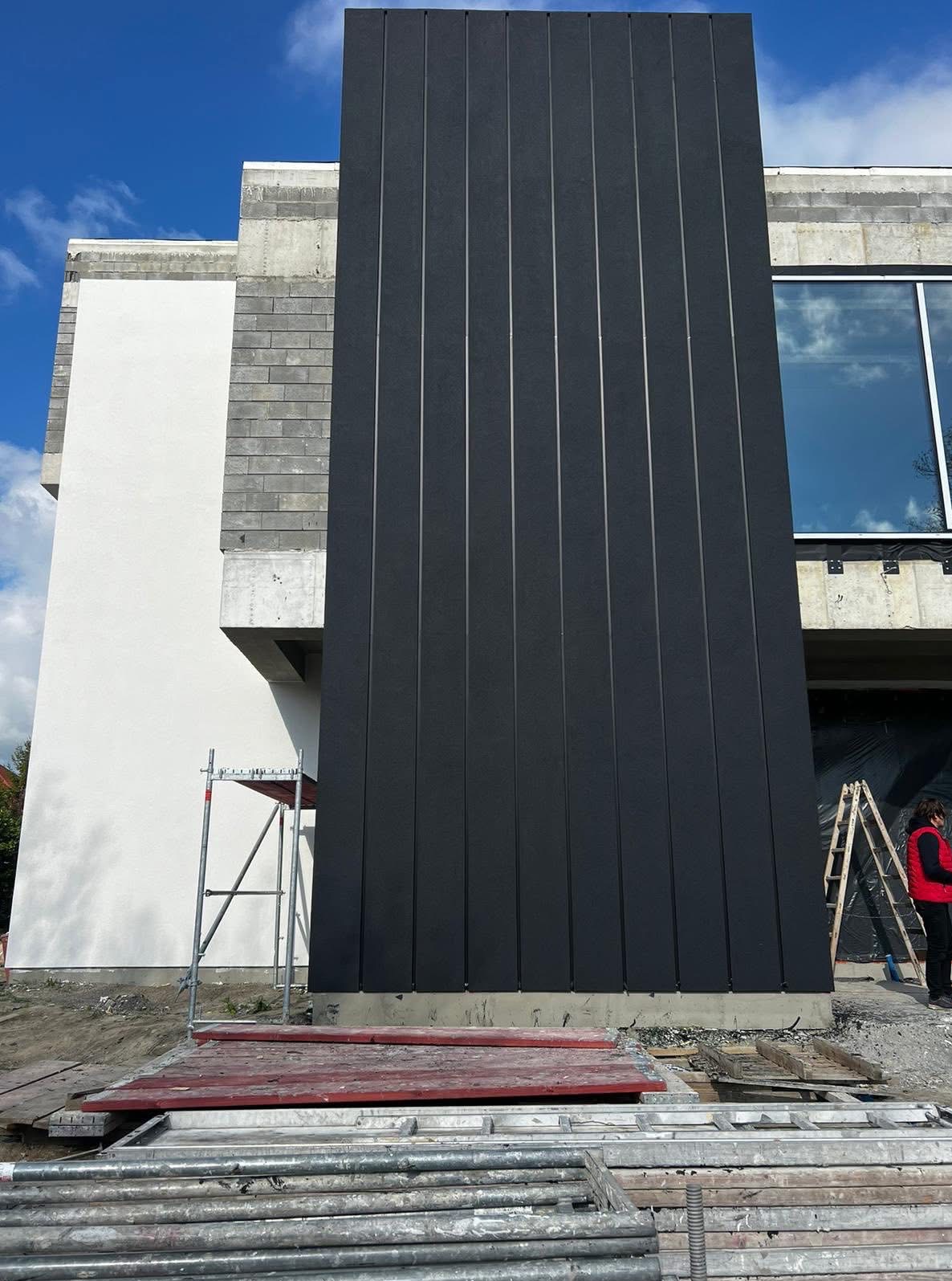 Nowoczesna elewacja budynku w trakcie wykończenia, z czarnymi panelami pionowymi. Widoczne rusztowania i elementy konstrukcyjne, okno z odbiciem nieba.