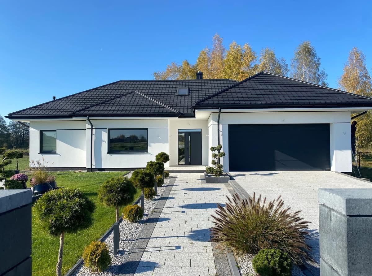 Nowoczesny dom jednorodzinny z czarnym dachem i garażem. Biała elewacja, szara kostka brukowa i ozdobne krzewy. Błękitne niebo w tle. Ujęcie z poziomu gruntu.