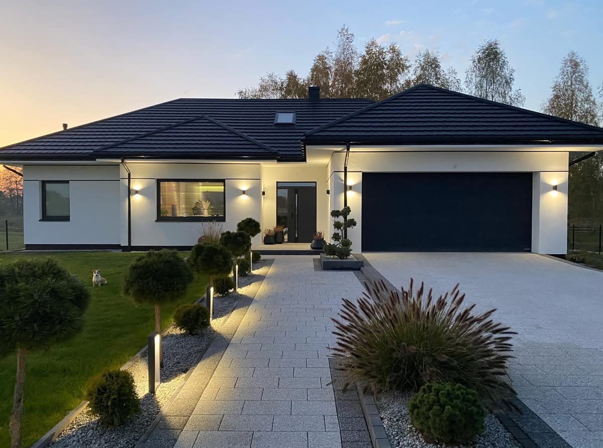 Nowoczesny dom jednorodzinny z czarnym dachem i garażem, oświetlony lampami. Na trawniku siedzi pies rasy Jack Russell Terrier. Kostka brukowa prowadzi do drzwi.